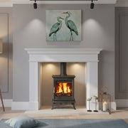 A modern living room scene featuring a black wood-burning stove insert in a white fireplace mantel, with a framed painting of two herons above. The room has gray walls, light wood floors, a floor lamp, and a blue rug.