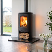 A black wood-burning stove with an open fire and visible logs, placed on a black base in a modern living room with large windows, a yellow chair, and a potted plant.