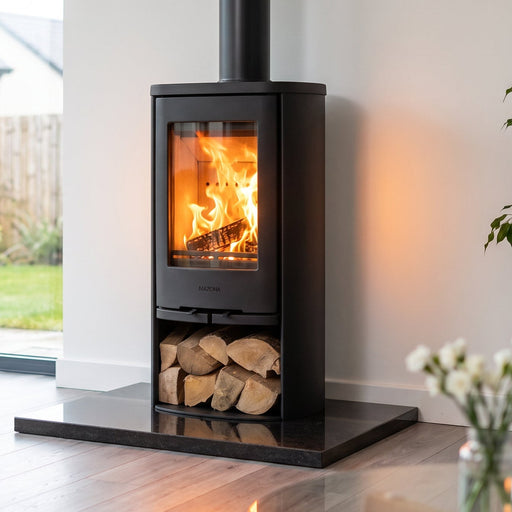 A black wood-burning stove with an open fire and visible logs, placed on a black base in a modern living room with large windows, a yellow chair, and a potted plant.