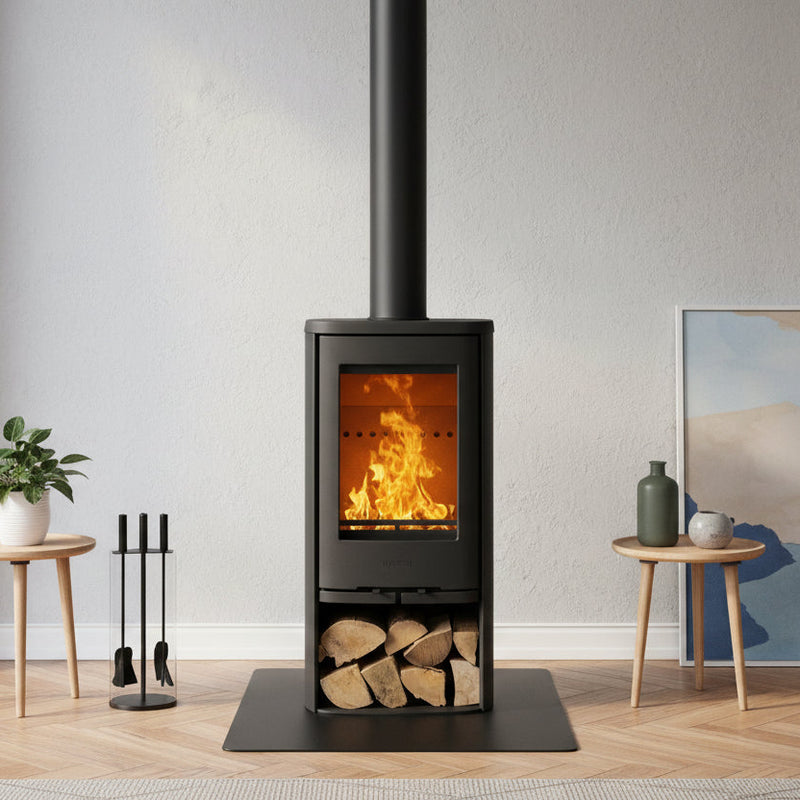 A black wood-burning stove with an open fire and visible flames, set in a modern living room with a light gray wall, wood flooring, and decorative side tables.