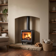 A black wood-burning stove with a glowing fire inside, set in a stone fireplace niche within a beige wall. The scene is styled with a stone hearth, stacked firewood, a woven basket of decorative spheres, and built-in wooden shelves with books and plants on both sides.