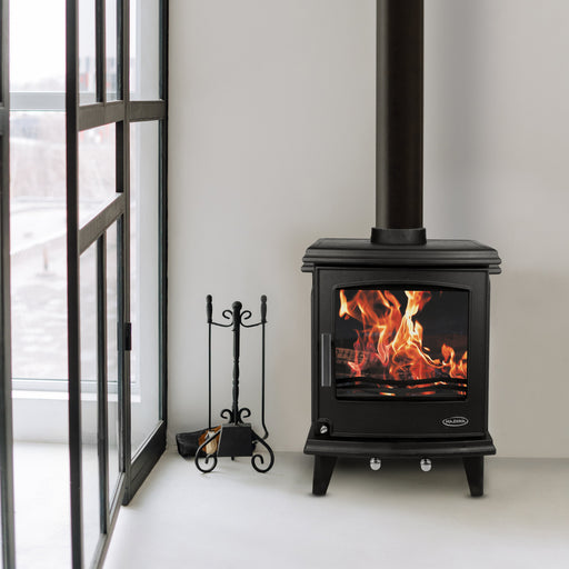 Black freestanding stove with flames inside, placed against a white wall next to a large window.